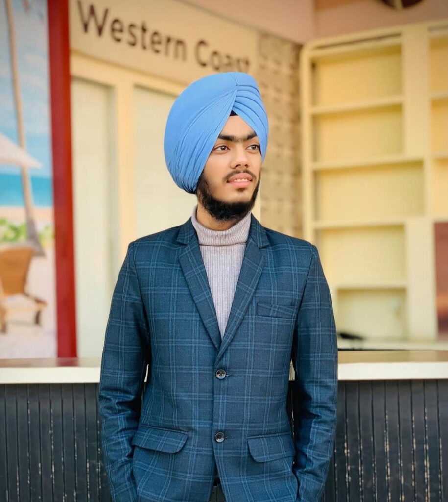 A young man wearing a blue turban and a tailored dark suit stands confidently, with a picturesque backdrop featuring a sign that reads 'Western Coast.'